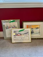 Three framed paintings resting against a wall, showing various winter rural landscapes