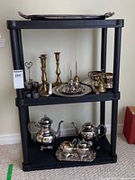 Full view of three-tier black shelf holding various silver-plated tea and coffee set items on the bottom shelf, brass candlesticks and silver trays on the middle shelf, and additional decorative items on the top shelf