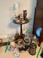 Small round wooden pedestal table with a dark finish holding decorative items including a glass hurricane vase with artificial flowers, tall wooden vase, small black birds, and candle holders. Additional decorative jars and glass vases surround the base.