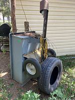 Side view of gray metal yard cart with two large tires and one smaller tire mounted on the side. Vertical brown post with yellow cable wrapped around it. Visible rust and wear on the metal surfaces. The cart is upright and appears to have dirt inside.