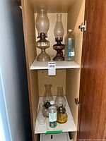 Two shelves inside a cupboard with four vintage glass oil lamps and four partial kerosene or lamp oil bottles.