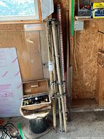 Vintage surveying level with tripod and grade rod positioned next to window and wall in workshop, showing case, tripod folded, and grade rod leaning against wall.