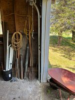 Photo of garden tools including shovels, pitchforks, rakes with wooden handles, metal pipes and extension cords hanging coiled on wall.