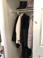 Photo shows several women's coats hanging in a closet: black trench coat, cream/white coat, light pink puffer jacket, and another dark coat. Above the coats are black boots and a pink baseball cap.