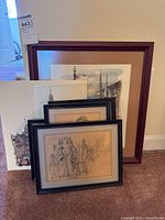 View of six pieces of artwork, including various framed prints leaning against a wall on a carpeted floor. Visible are pencil sketch portraits and architectural watercolor prints with different frame styles.