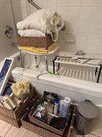 Photo showing white bathrobes folded on woven basket on top of a white plastic corner shelf with folded towels underneath. Three woven baskets below containing personal care products like deodorant, razor, and other items. Silver metal pedal garbage can next to baskets on tiled floor.