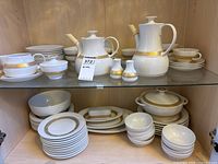 Full set displayed inside cabinet showing cups, saucers, plates, bowls, teapots, creamer, sugar bowl, and salt and pepper shakers with matching gold trim.