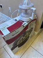 Rival electric wok in original box with electric juicer on top near wall outlet.