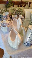 Overall display of fine china tea set including teapot, cups, saucers, and swan-shaped porcelain vases on a white lace tablecloth.
