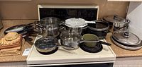 Wide view of assorted pots, pans, colander, double boiler arranged on white stove.