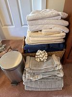 Woven basket holding multiple towels with additional folded towels stacked in front and underneath, alongside small silver garbage can.