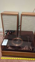Turntable with transparent dark brown dust cover and two wooden speakers with fabric fronts and attached cables.