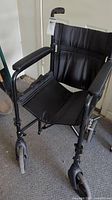 Full frontal view of black lightweight wheelchair with padded armrests, fabric seat and back, and front caster wheels.