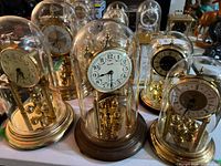 Grouped photo of five anniversary clocks under glass domes showing various dial styles and brass tone metal parts.