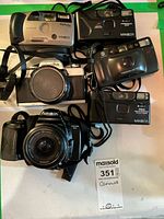 All six Minolta cameras pictured together on a table, showing the variety of models including point & shoot and SLR cameras with different lens types and colors.