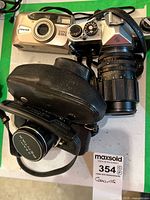 Lot of two Pentax 35mm SLR cameras and one Pentax Espio 838S point and shoot camera shown together on a white surface with green background. One SLR is in a black leather carrying case.