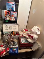 Lot contents arranged on a surface showing teddy bear, collector plates, ceramic teapot and mugs, Sylvania Christmas lights boxes, wooden box with winter scene, figurines and coasters.