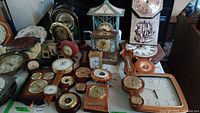 Overall view showing an assortment of clocks and barometers on a table with varied styles and sizes from 2.5 to 16 inches, including wooden, brass, and ceramic pieces.