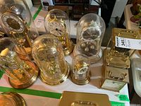 Overview of 7 clocks including 5 anniversary clocks under glass domes and 2 brass mantle clocks on tabletop.