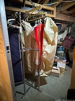 Front view of chrome finish industrial clothing rack with clothes hangers and garment bags hung on it, in a storage or basement setting.