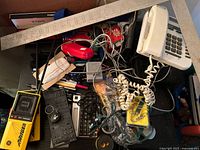 Desk surface showing disorganized mix of electronics and office supplies including white telephone, black keyboard, red device, cables, and boxed pens.