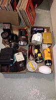 Photo showing several flashlights including black and yellow handheld types, a purple Conair hair dryer, and assorted wallets and cases in cardboard boxes on a concrete floor