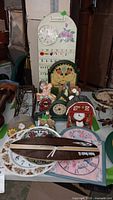 Photo of lot showing a variety of novelty clocks including a barn-shaped clock, floral plate clocks, and a tall calendar clock.