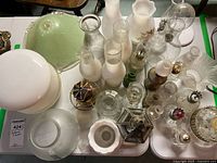 Overview of various glass lamp globes, shades, and oil lamp parts showing a mix of white, clear, and pale green glass pieces including chimneys and bases.