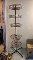 Full view of tall wire rack with five circular wire baskets mounted on a central pole and tripod base in a corner against a wall, surrounded by other stored items.