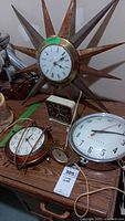 Photo shows five vintage clocks on a wood surface including the large sunburst wall clock with metal rays, two rectangular desk clocks, and two round wall clocks.