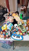 Table with various toddler toys and dolls including a toy piano keyboard, soccer ball, plush toys, and diecast vehicles in packaging
