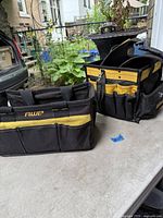Two tool bags on a concrete floor with garden and fence background, one DeWalt and one AWP