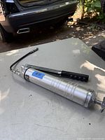 Metal grease lubricator with flexible hose and pump handle shown on a table outdoors near a vehicle.