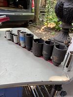 Photo showing a row of 9 impact sockets of different sizes on a red rail, with a silver adapter on the right.