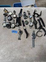 Photo shows 18 assorted watches laid out on a table, including pocket watches, wristwatches with metal bands, leather straps, and rubber bands, varying in style and condition.