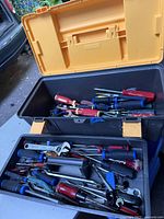 Open tool box with yellow lid showing various hand tools including multiple screwdrivers with red and blue handles, wrenches, and pliers inside.