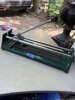 Green manual tile cutter shown from front side placed on a table outdoors, displaying the handle lever and cutting wheel on metal rails.