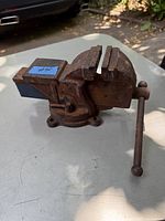 Photo showing a rusty vintage metal bench vise placed outdoors on a flat surface with greenery in the background.
