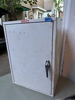Closed view of the white metal wall cabinet showing handle and some wear.