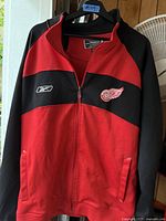 Front view of red and black Reebok NHL jacket with Detroit Red Wings logo on left chest and Reebok logo on right chest.
