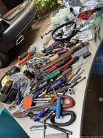 Overview of assorted hand tools and hardware laid out on a table including pliers, screwdrivers, clamps, hammers, sockets, and drill bits.