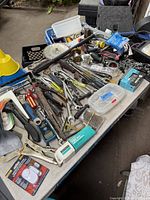 Overview of assorted hand tools, hardware, adhesive, measuring tools, and power tool accessories spread on a table. Includes wrenches, pliers, screwdrivers, a cordless drill, sealant tube, door alarms, paint scraper, and yellow funnel.