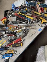 Top view of assorted hand tools and hardware items spread out on table, showing knives, levels, tape measures, ratchets, sockets, and screwdrivers.