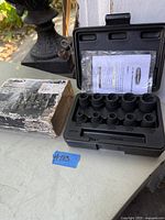 Contents of open black plastic case showing 10 socket nut extractors arranged in molded slots, instruction sheet inside, next to worn cardboard box