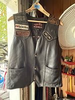 Front view of black vintage leather vest showing six sewn patches and two front pockets.