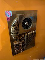 Full view of a brass or gold-tone metal wall art sculpture with a turbine circular centerpiece, crescent shape with cutouts on left, and ribbed rectangular elements at bottom right, all mounted on a rectangular backing.