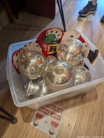Clear photo of five mercury glass Christmas ornaments and a Santa Claus plate inside a clear plastic bin on a wood floor next to furniture.