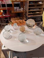 Overview of all eight glass art paperweights arranged on a round white table showing variety in shape and bubble designs including a grape cluster shaped piece in peach glass.