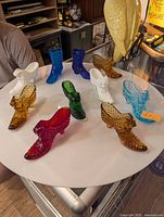 Nine glass boots in various colors (white, blue, amber, green, red, aqua) displayed on a white round table indoors, featuring hobnail texture.