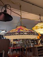 Photo of pendant light featuring a wide round shade decorated with multicolor beadwork and geometric patterns, hanging by a gold chain from a dark ceiling canopy in a shop setting.